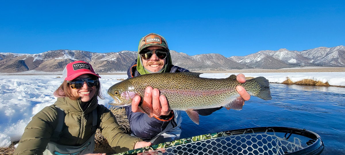 A smiling fly fisherman holding a rainbow trout on a river.