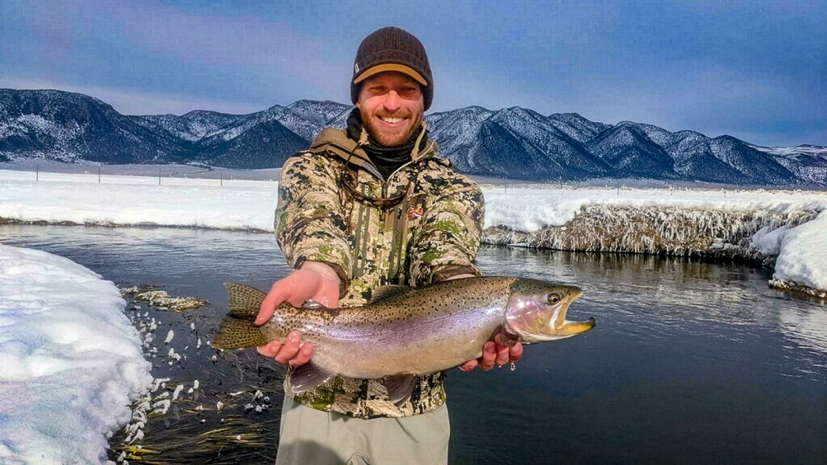 A smiling fly fisherman holding a rainbow trout on a river.