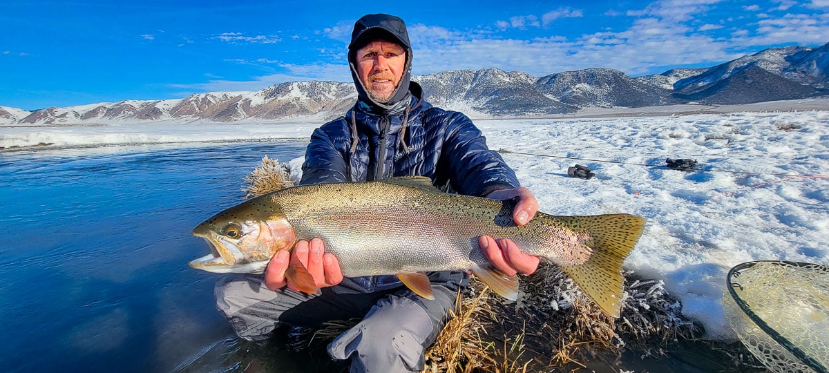 A smiling angler holding a giant rainbow trout on a river.