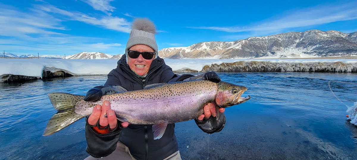 A smiling female angler holding a giant rainbow trout on a river.