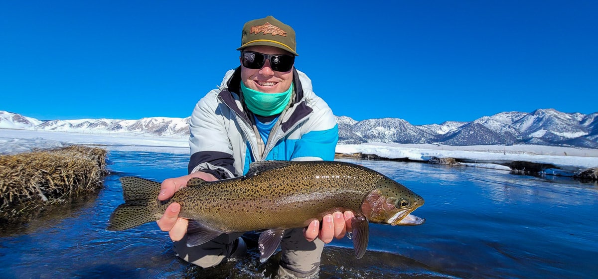 A smiling fly fisherman holding a rainbow trout on a river.