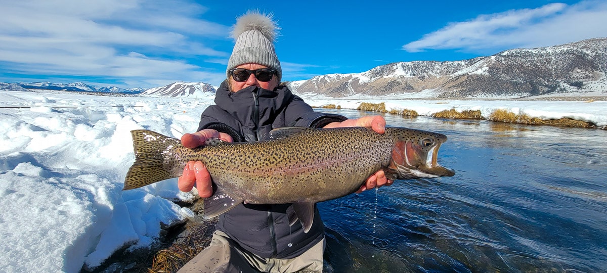 A smiling female angler holding a giant rainbow trout on a river.