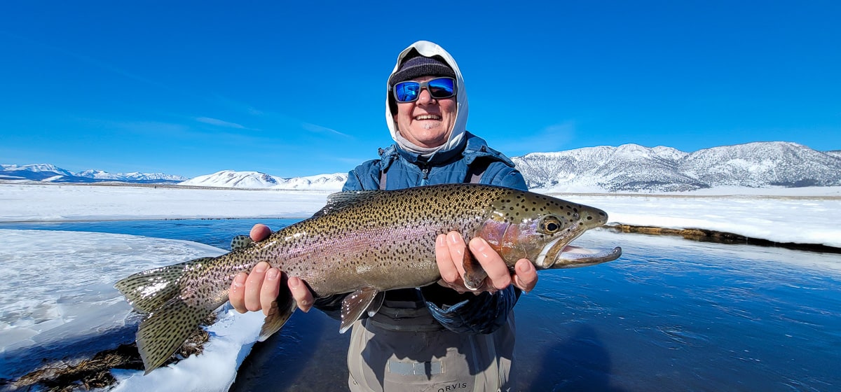 A smiling fly fisherman holding a rainbow trout on a river.