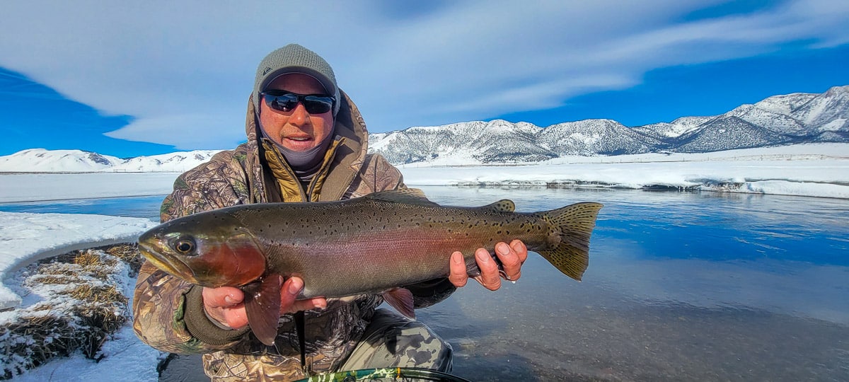 A smiling fly fisherman holding a rainbow trout on a river.