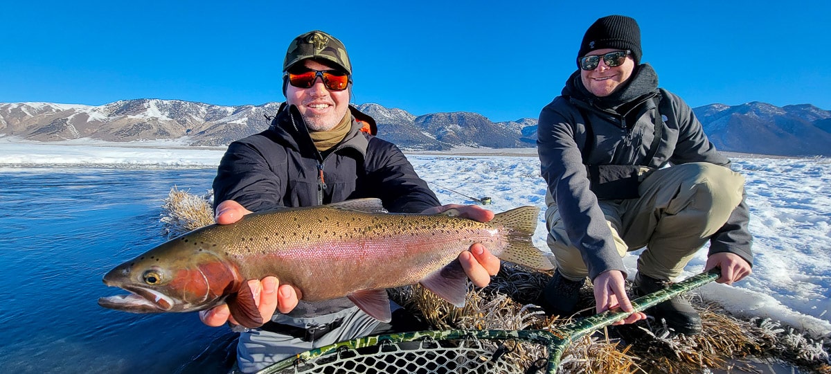 A smiling fly fisherman holding a rainbow trout on a river.