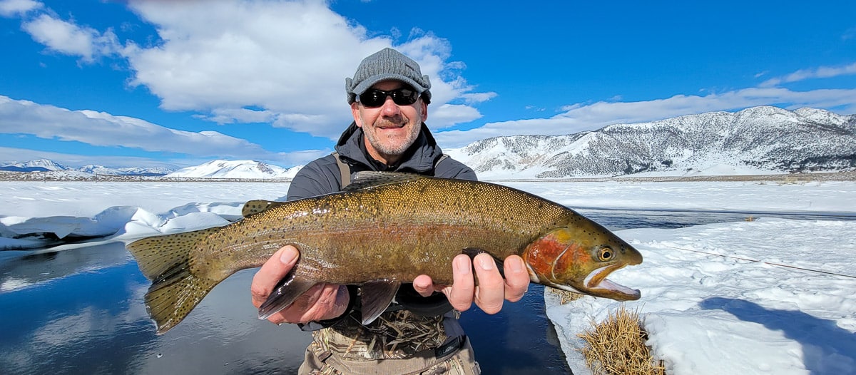 A smiling fly fisherman holding a rainbow trout on a river.