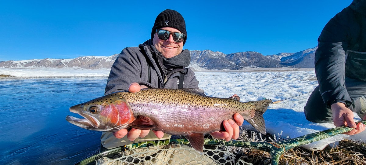 A smiling fly fisherman holding a rainbow trout on a river.