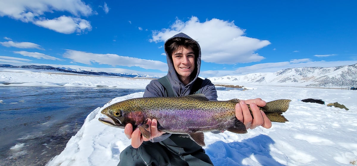 A smiling fly fisherman holding a rainbow trout on a river.