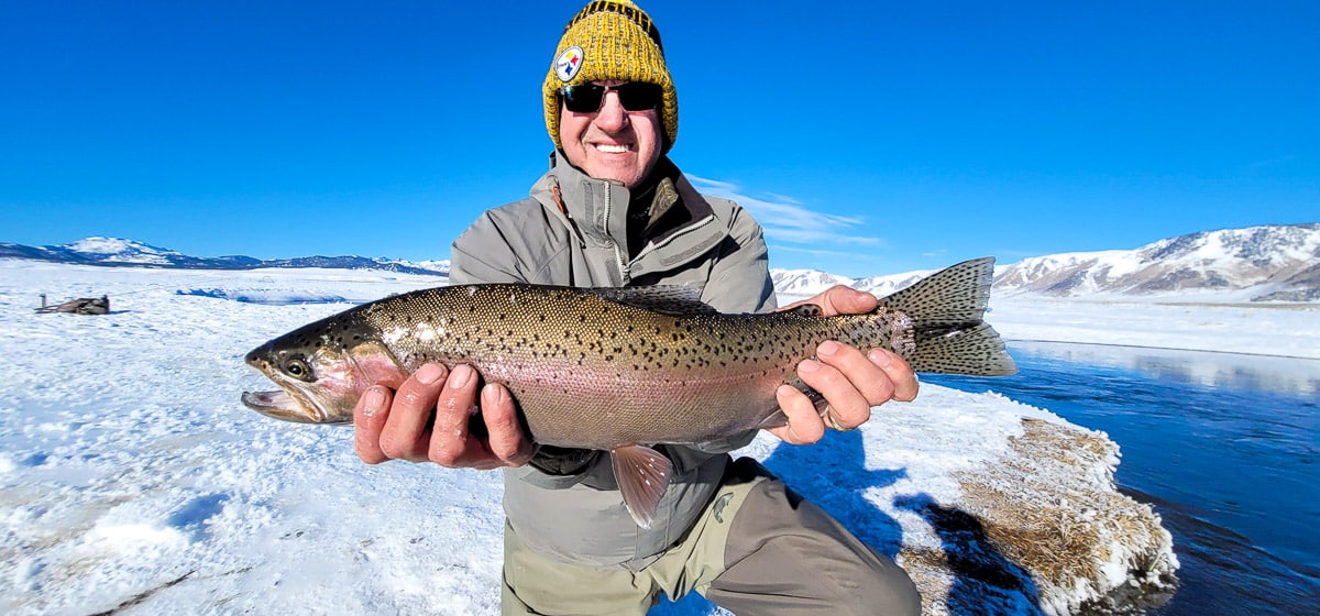 A smiling fly fisherman holding a rainbow trout on a river.