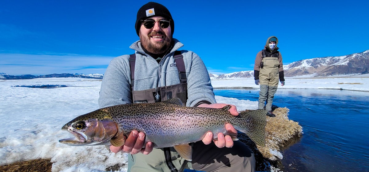 A smiling fly fisherman holding a rainbow trout on a river.