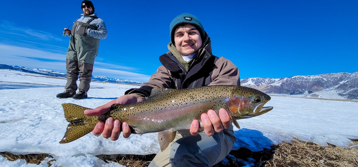 A smiling fly fisherman holding a rainbow trout on a river.