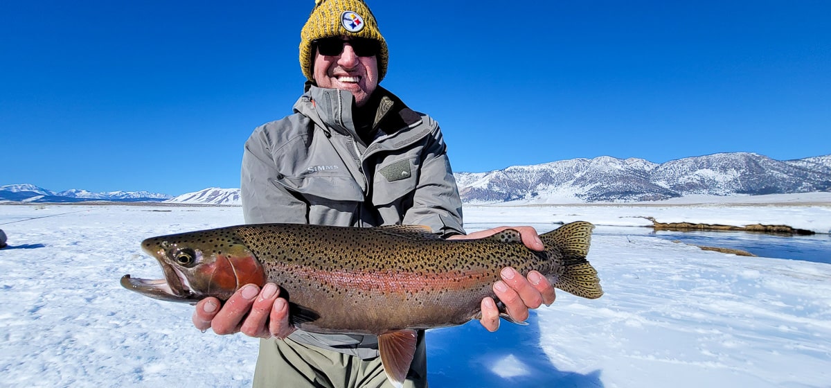 A smiling fly fisherman holding a rainbow trout on a river.