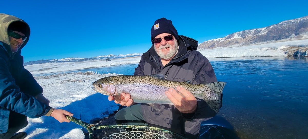 A smiling fly fisherman holding a rainbow trout on a river.