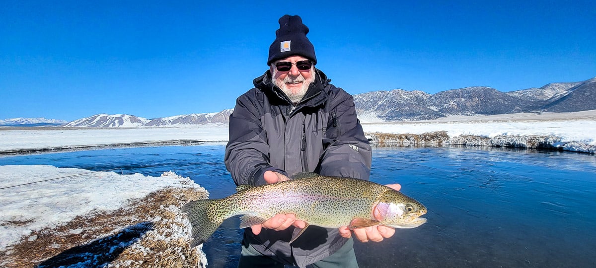 A smiling fly fisherman holding a rainbow trout on a river.