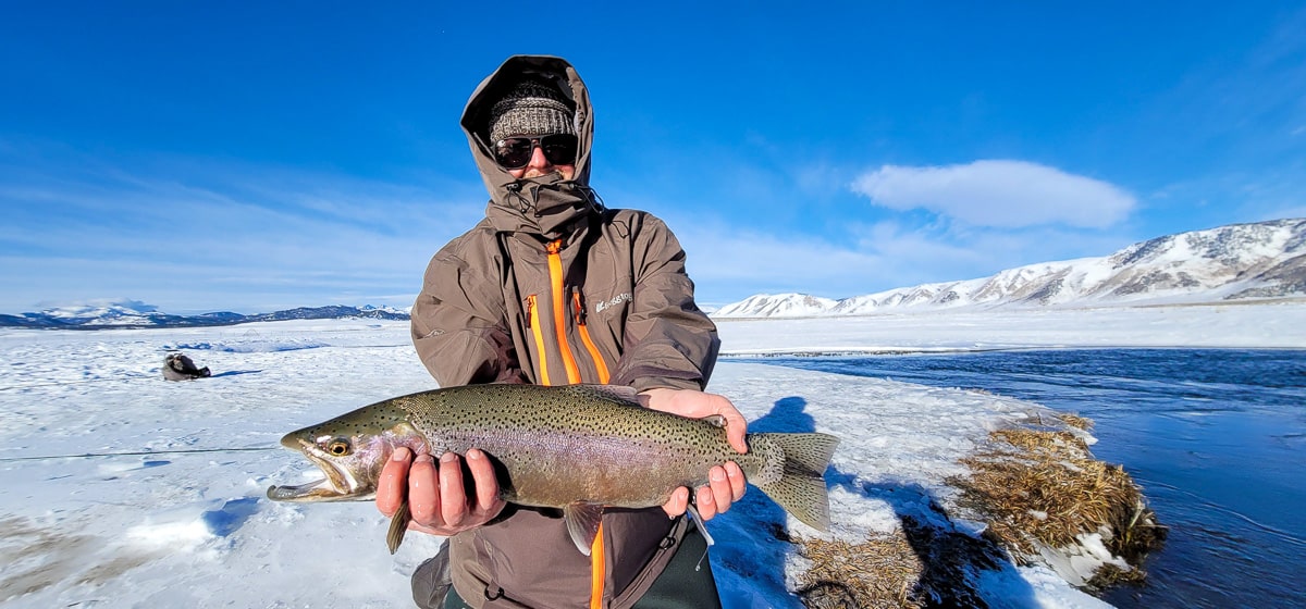 A smiling fly fisherman holding a rainbow trout on a river.