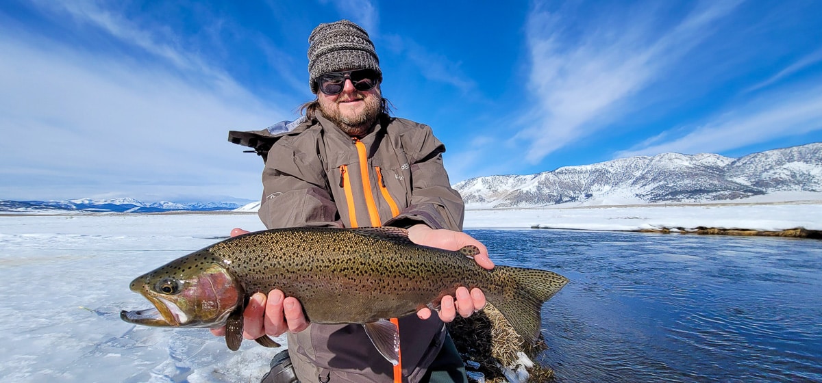 A smiling angler holding a giant rainbow trout on a river.