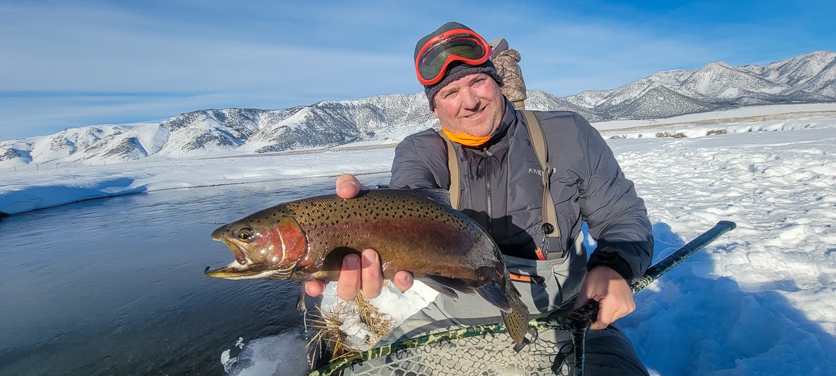 A smiling fly fisherman holding a rainbow trout on a river.