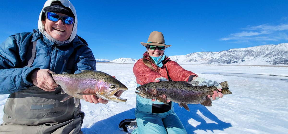 A man and woman angler fishing in the winter and holding a pair of giant rainbow trout in the snow.