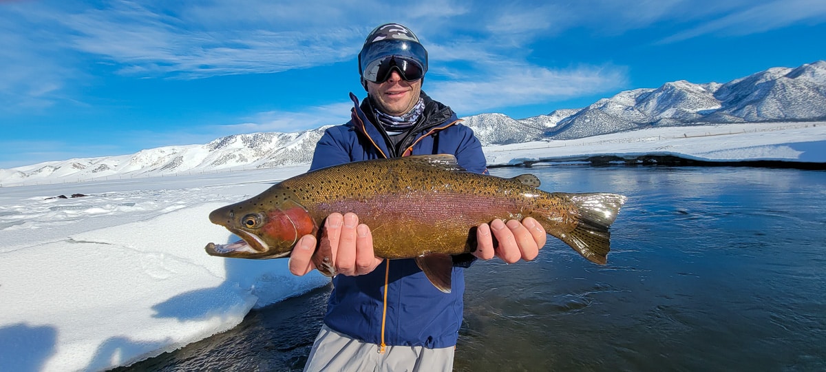 A smiling fly fisherman holding a rainbow trout on a river.