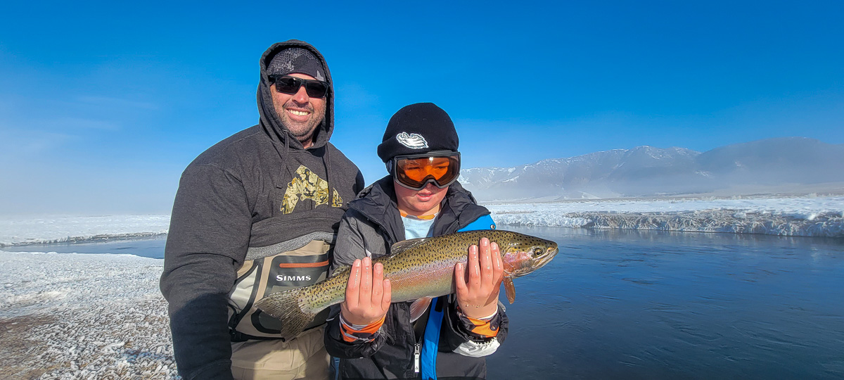 A man and a young boy display a larger rainbow trout with snow covered ground in the background and a river flowing around them.