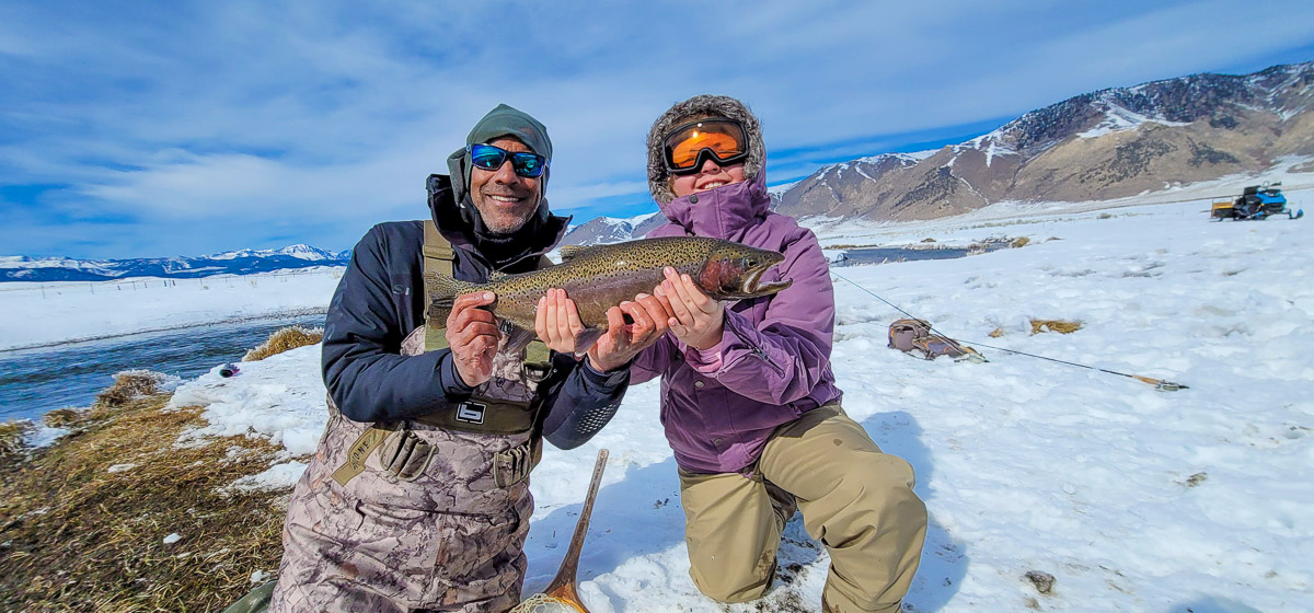 A man and a young female angler holding a larger rainbow trout on a snowy river.