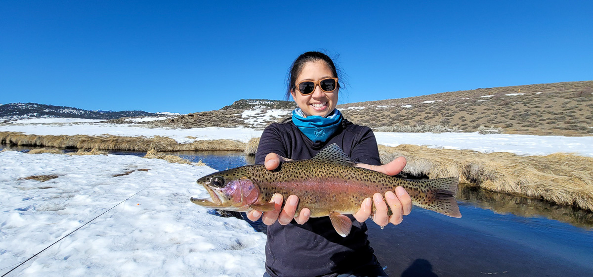 A smiling lady fly fisherman holding a rainbow trout on a river.