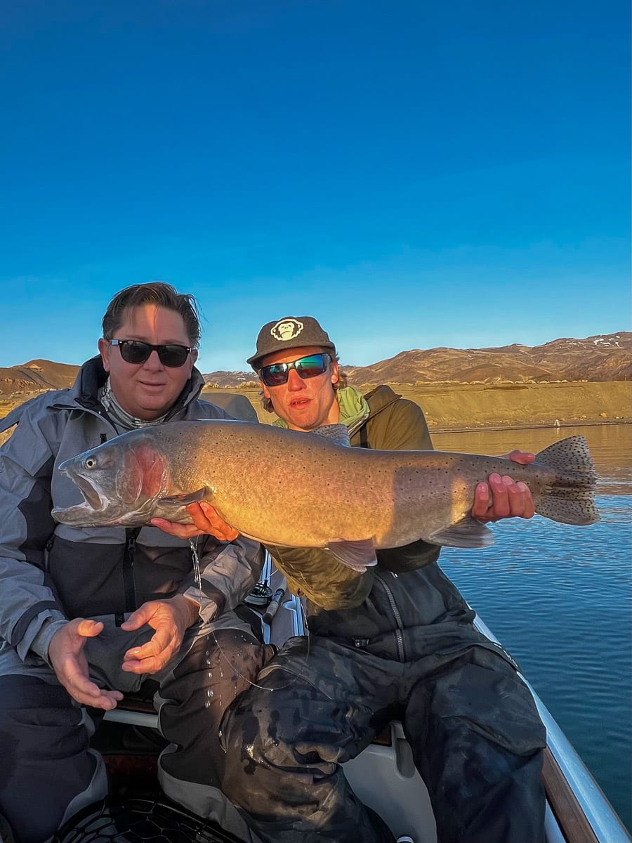 A smiling fly fisherman holding a giant cutthroat trout on a lake.