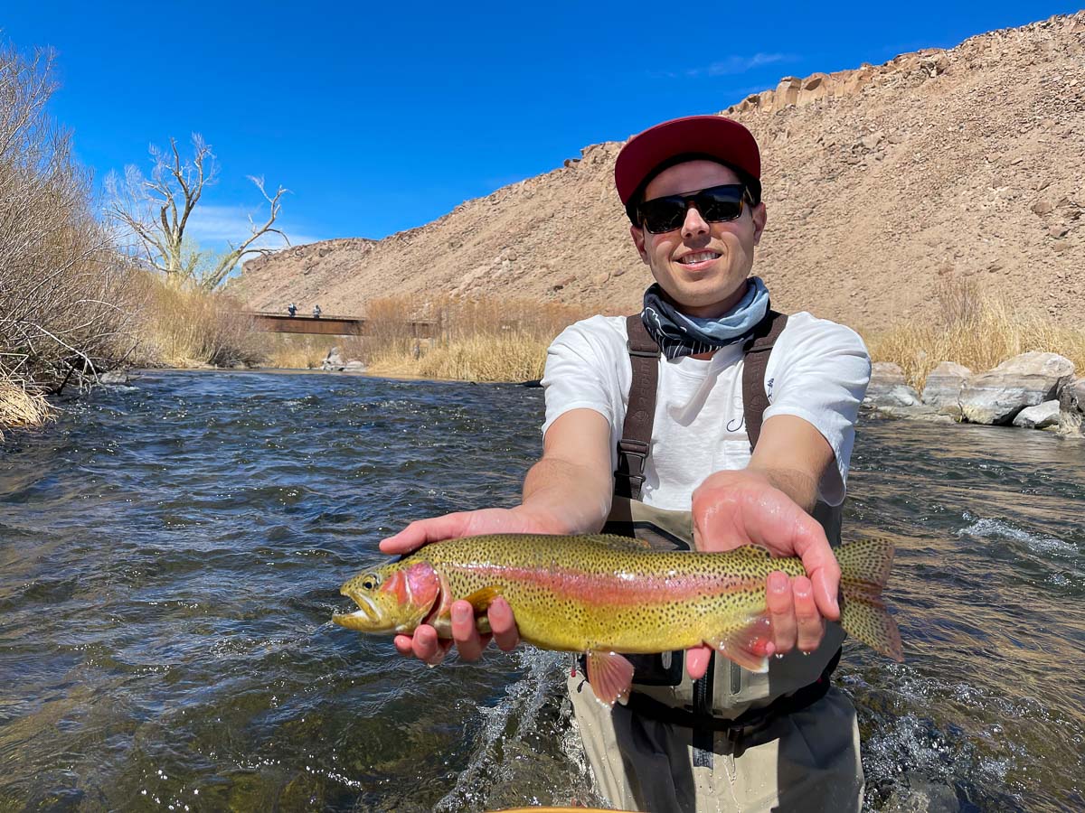 A smiling angler holding a rainbow trout on a river.