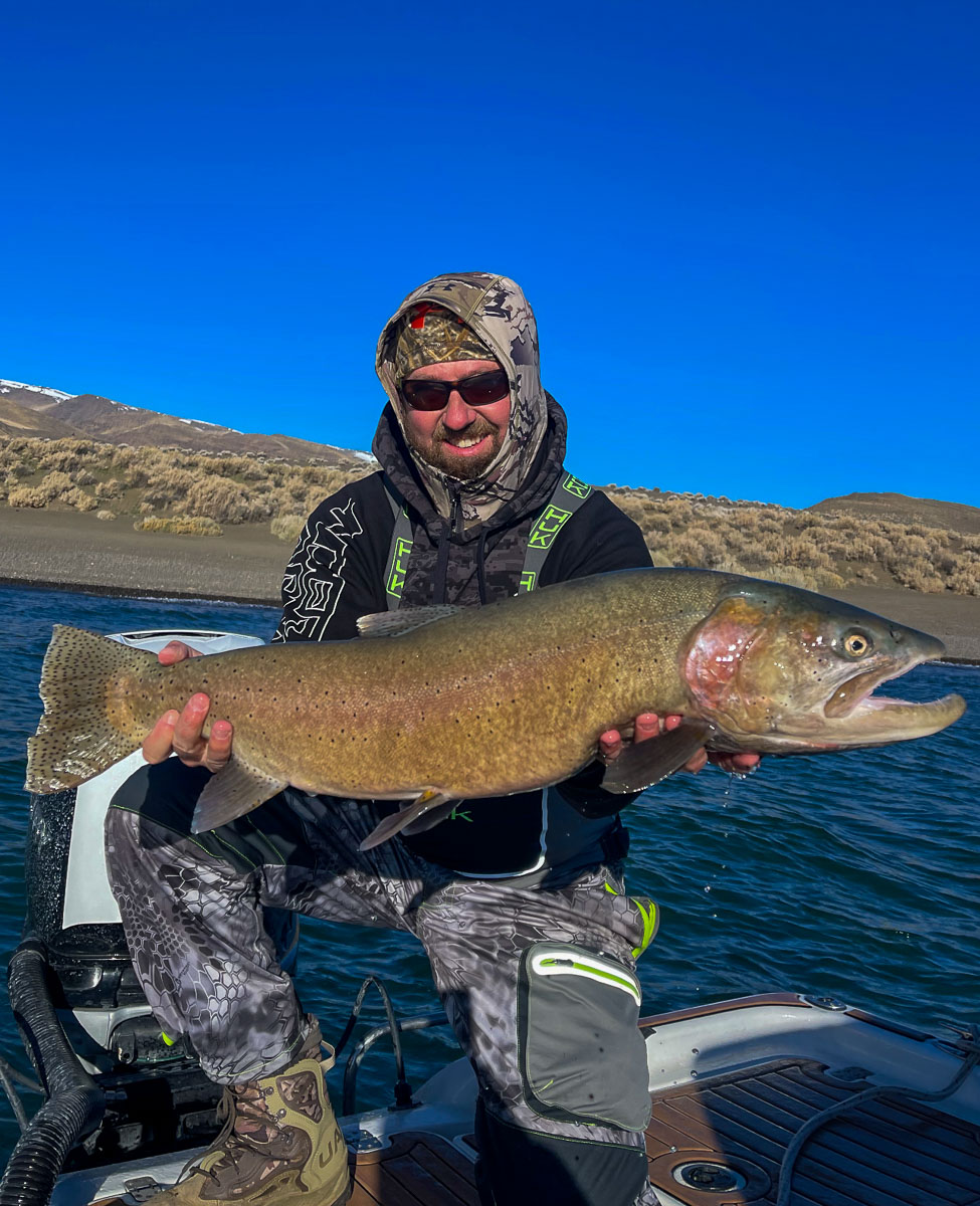 A fly fisherman on a lake in a boat holding a behemoth cutthroat trout.