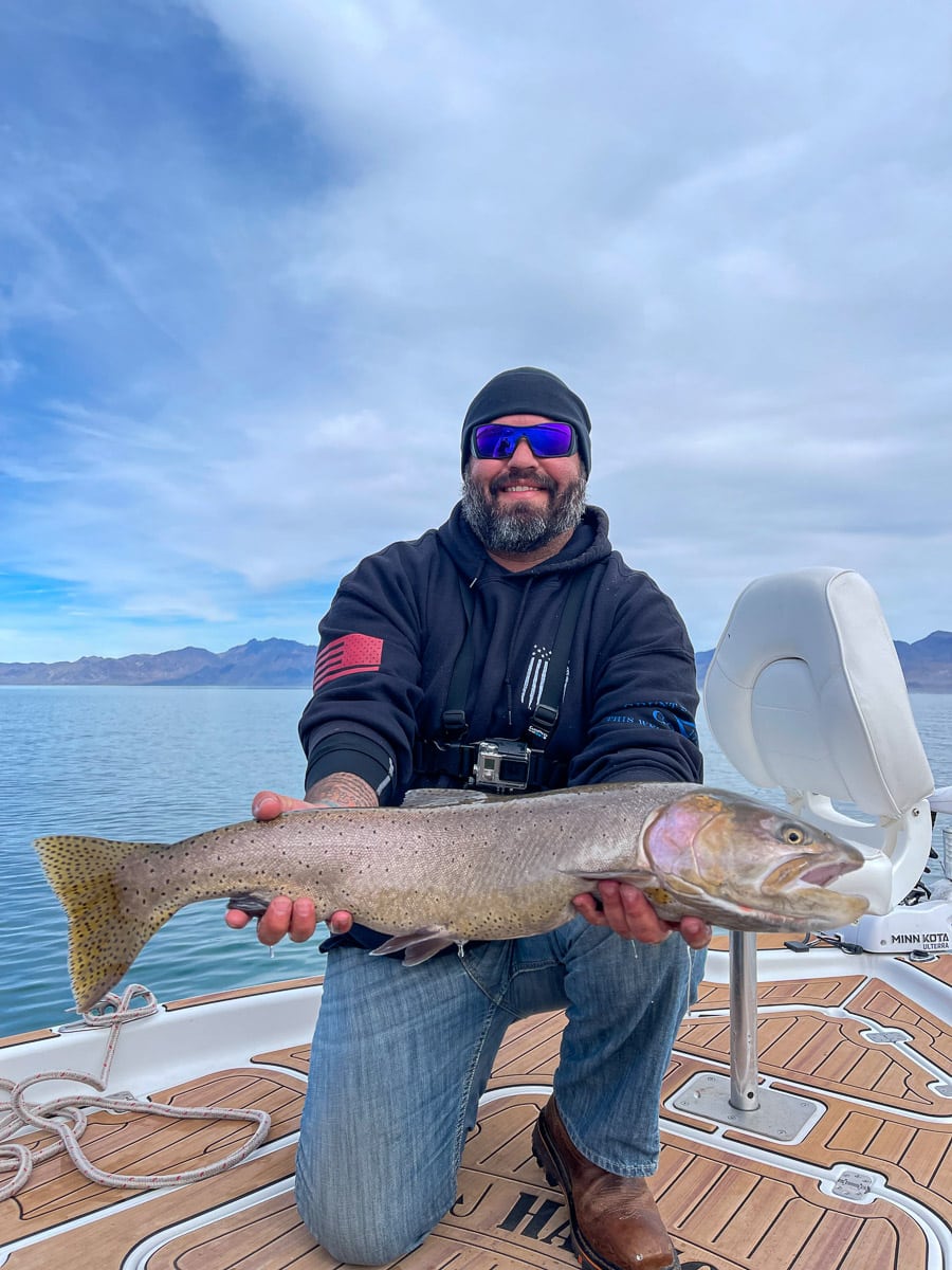 A fly fisherman on a lake in a boat holding a behemoth cutthroat trout.