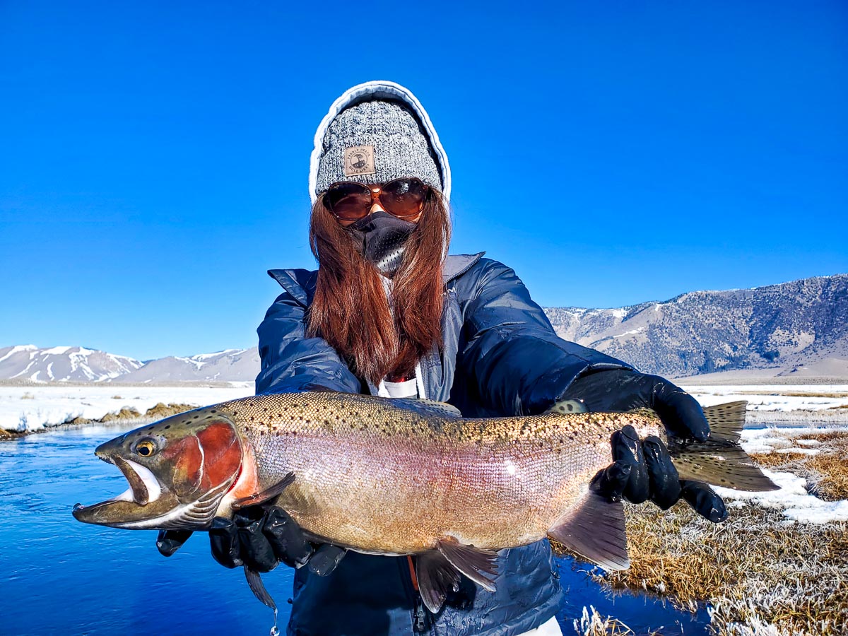 A smiling female fly fisherman holding a rainbow trout on a river.