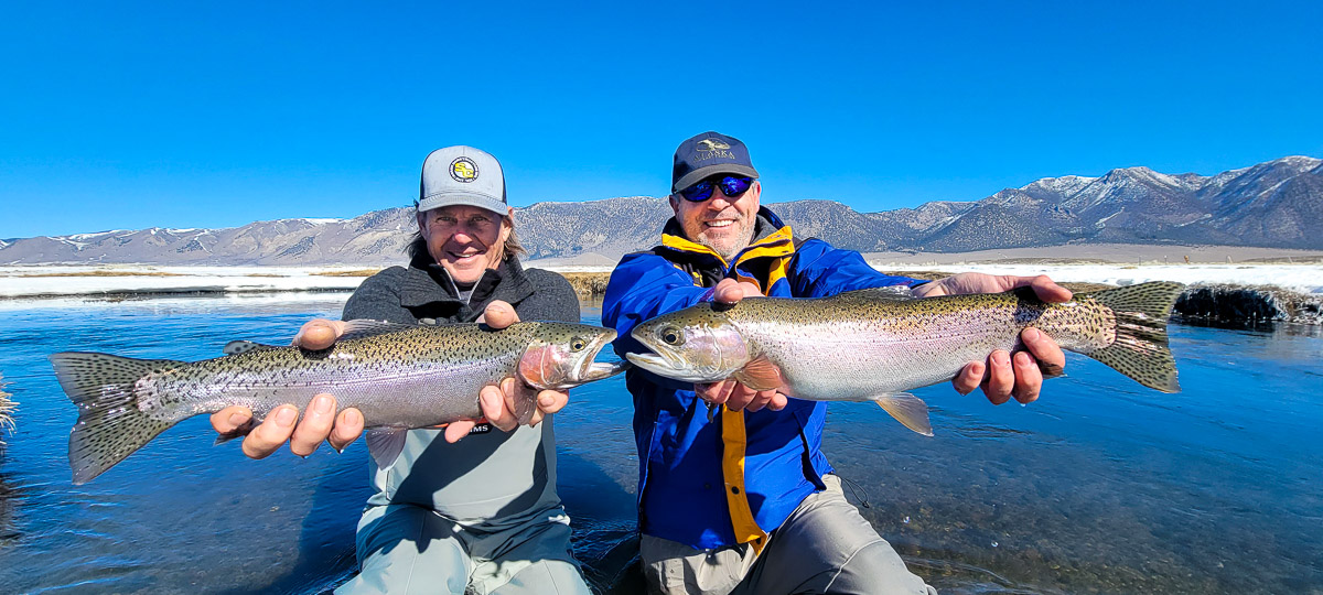 A smiling pair of fly fisherman holding a rainbow trout on a river.
