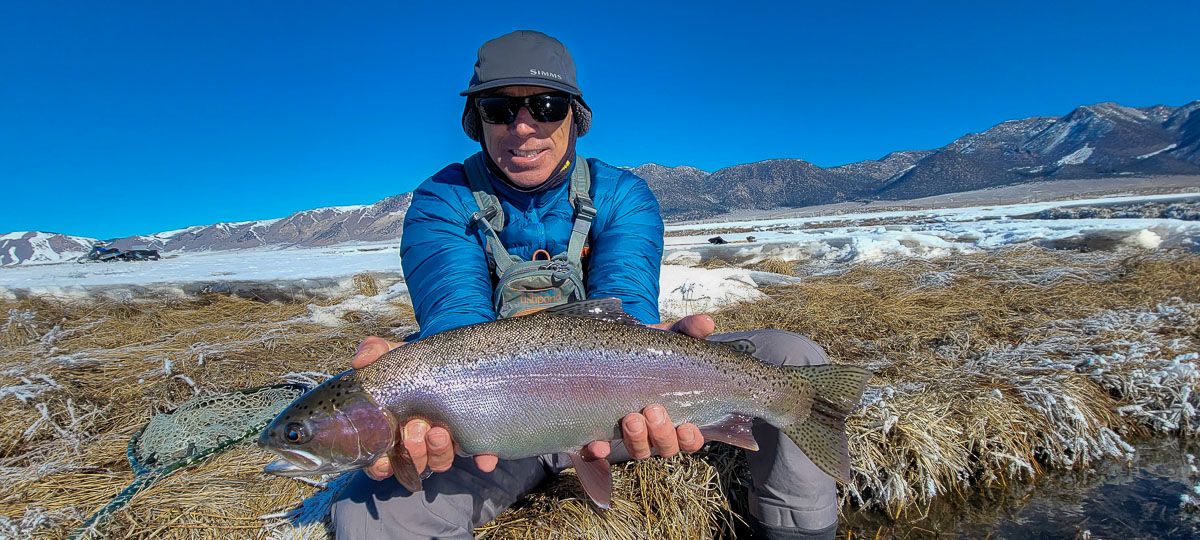 A smiling angler holding a giant rainbow trout on a river.