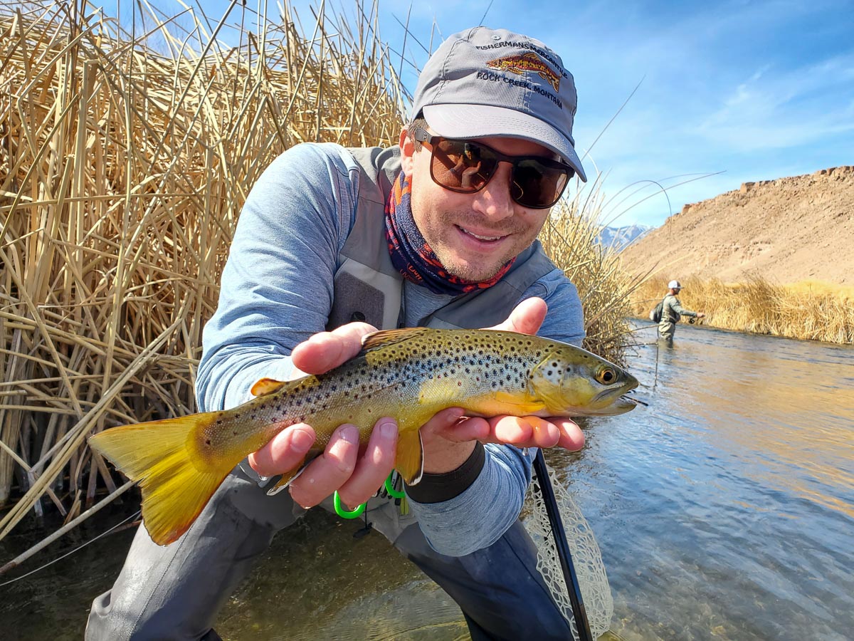 A smiling fly fisherman holding a brown trout on a river.