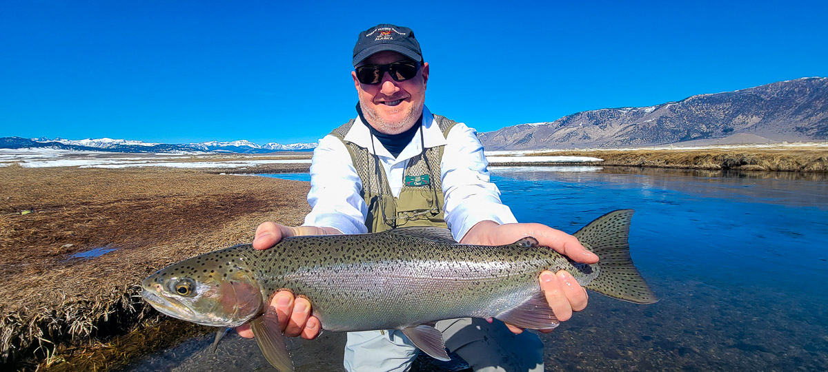 A smiling angler holding a giant rainbow trout on a river.