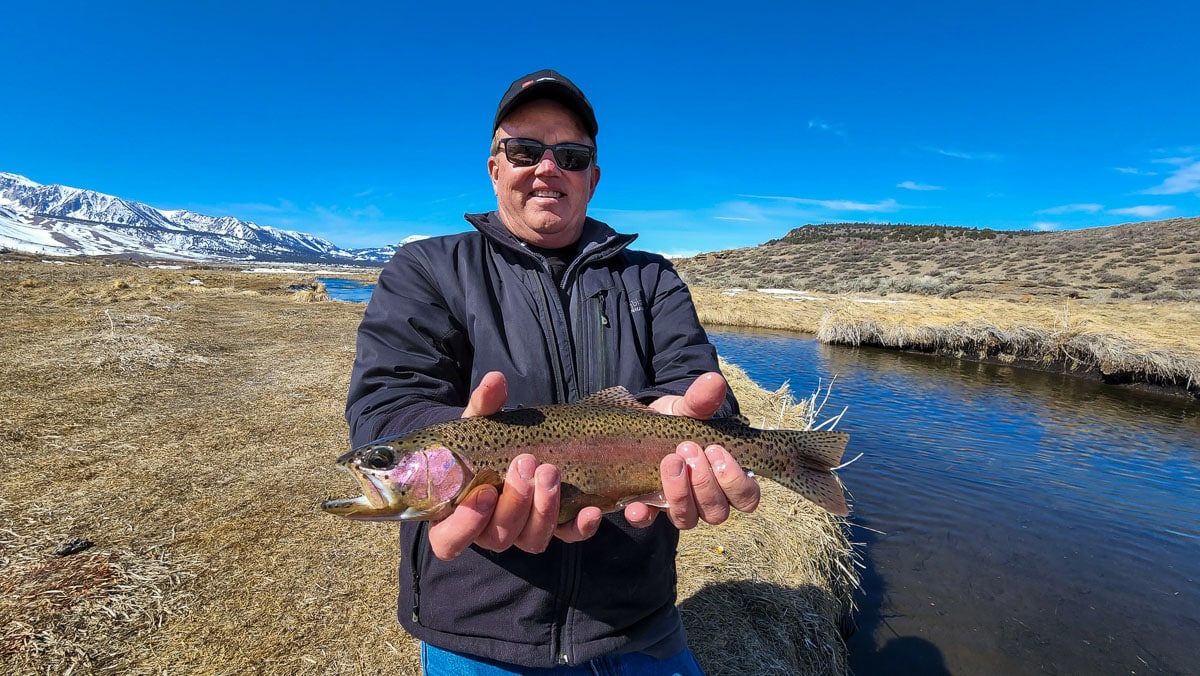 A smiling angler holding a giant rainbow trout on a river.