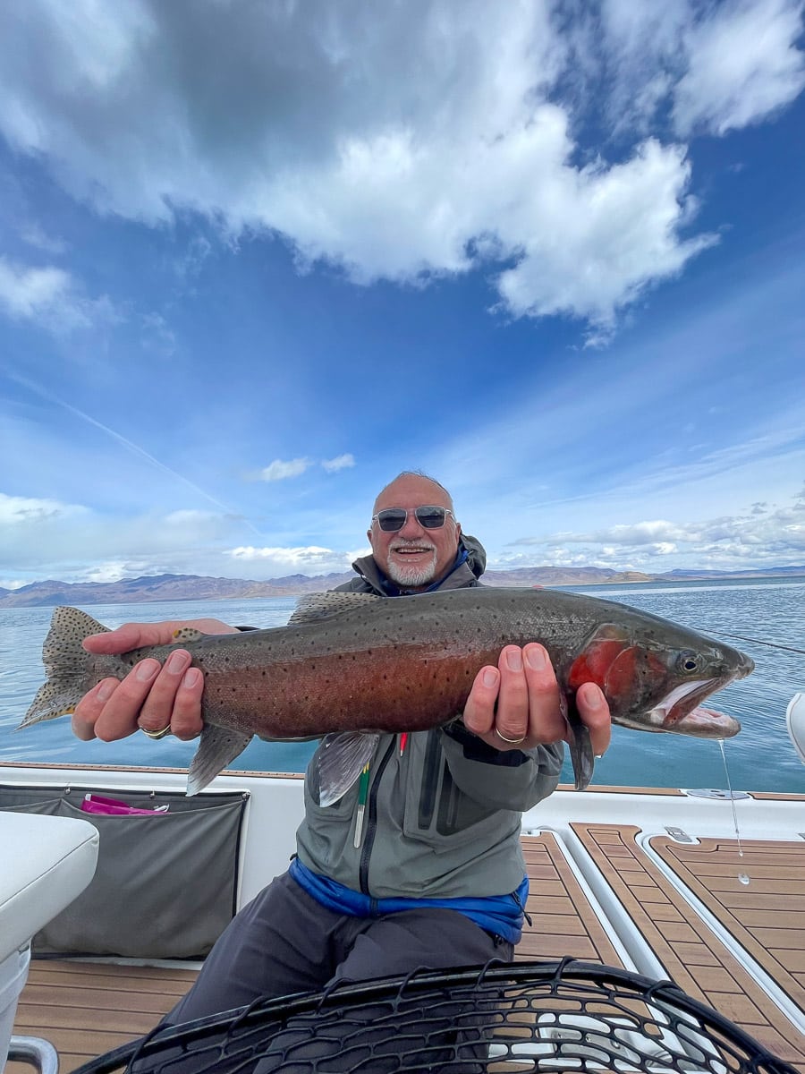 A fly fisherman on a lake in a boat holding a behemoth cutthroat trout.