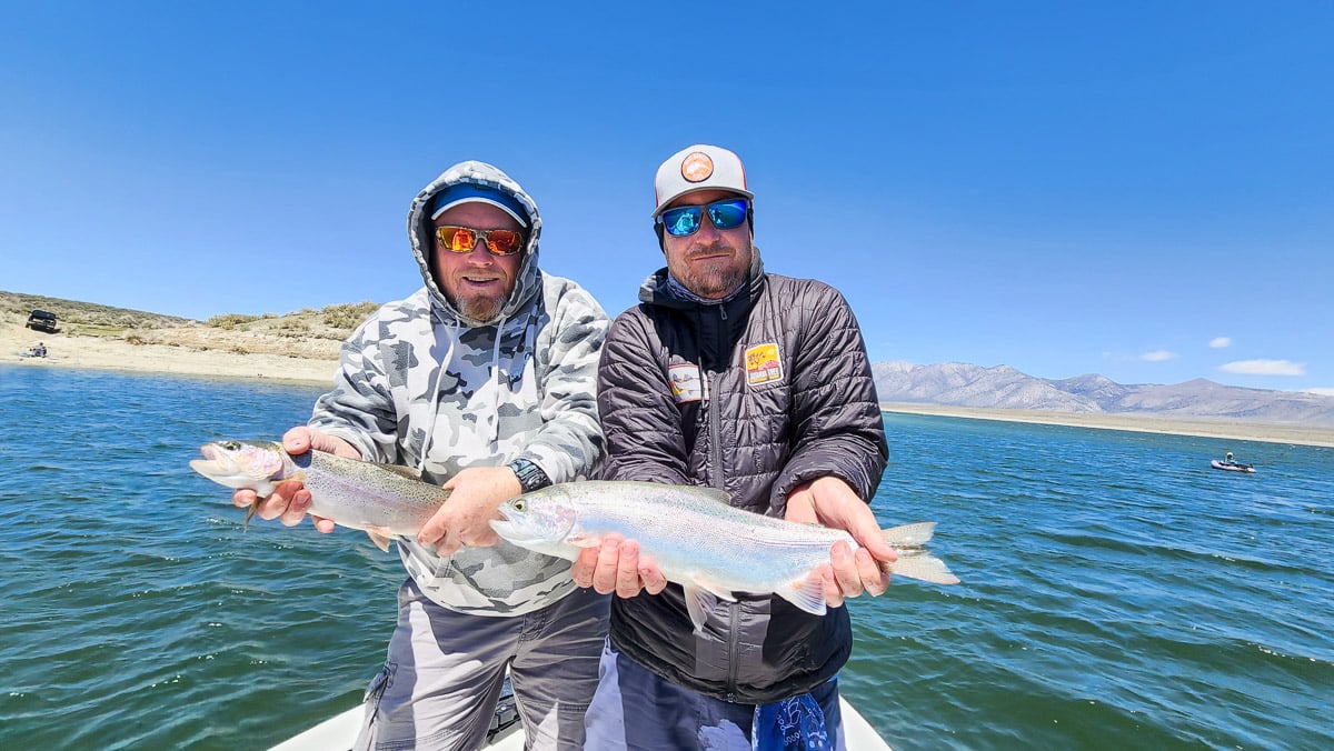 A pair of fishermen in a boat on a lake holding a pair of large rainbow trout.