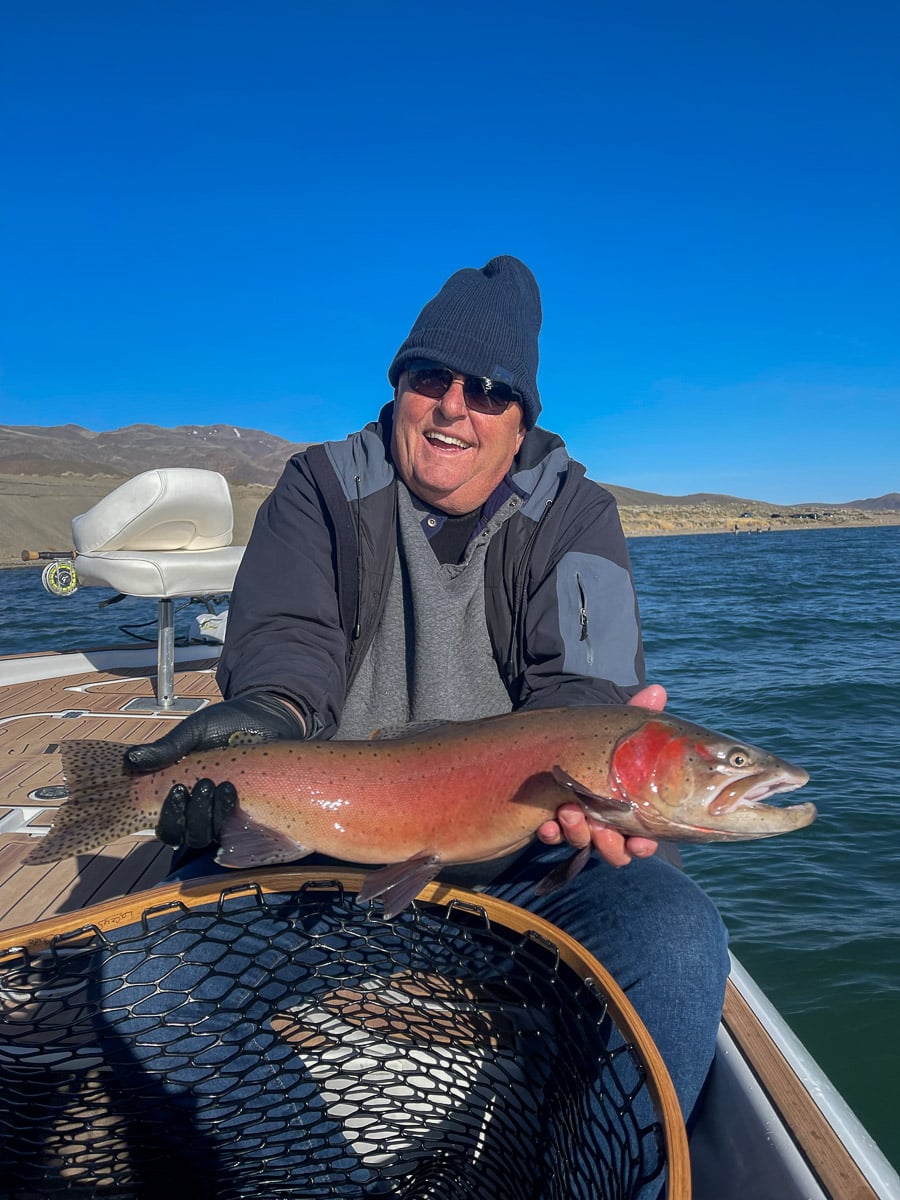 A fly fisherman on a lake in a boat holding a behemoth cutthroat trout.