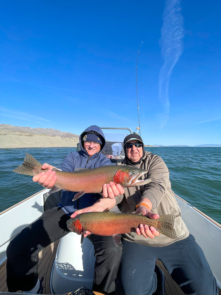 A pair of fishermen in a boat on a lake holding a pair of large cutthroat trout.