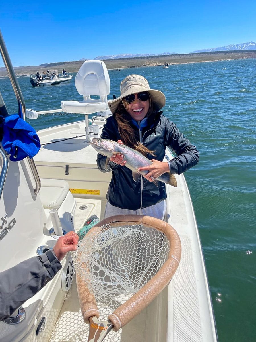 A smiling lady fly fisher on a lake in a boat holding a large rainbow trout.