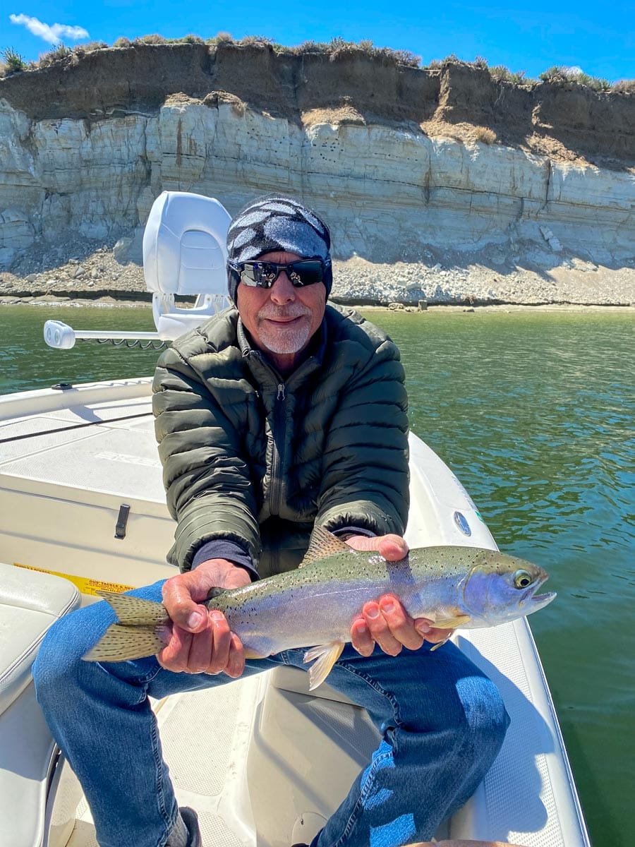 A fly fisherman on a lake in a boat holding a behemoth rainbow trout.