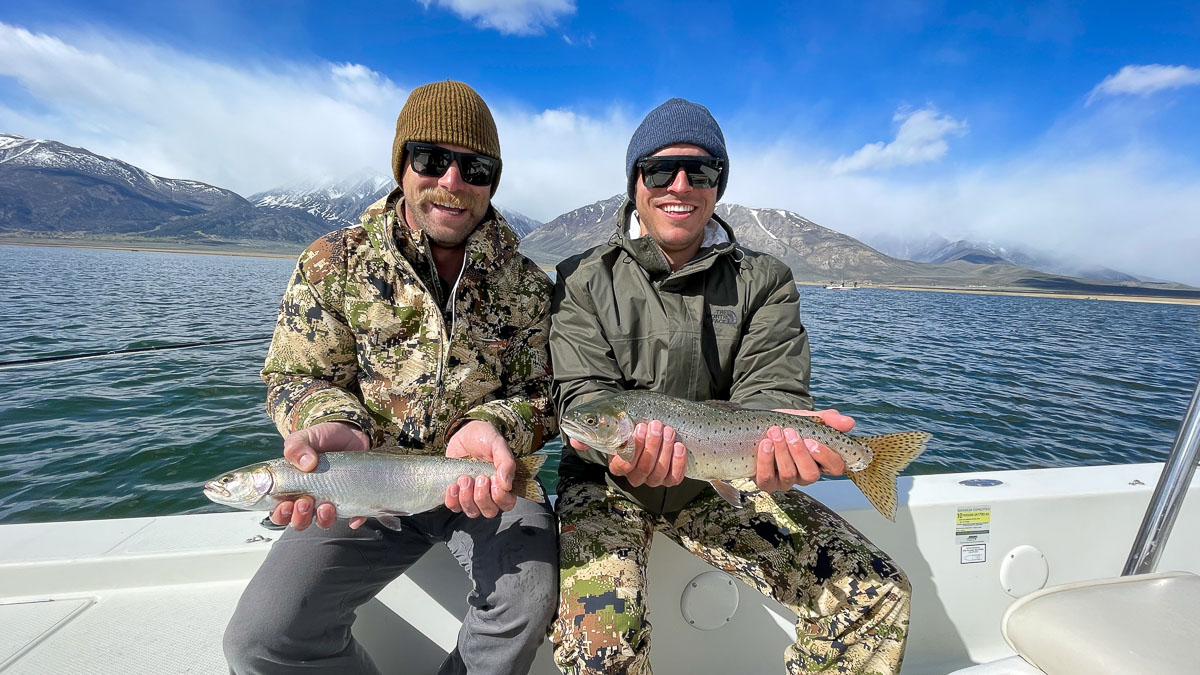 A couple of anglers dressed in camouflage clothing and beanie hats holding a pair of cutthroat trout on a lake.