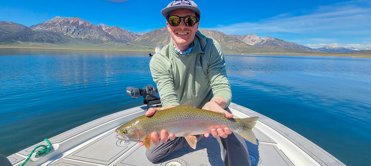 A smiling angler holding a large rainbow trout in a boat on a lake.