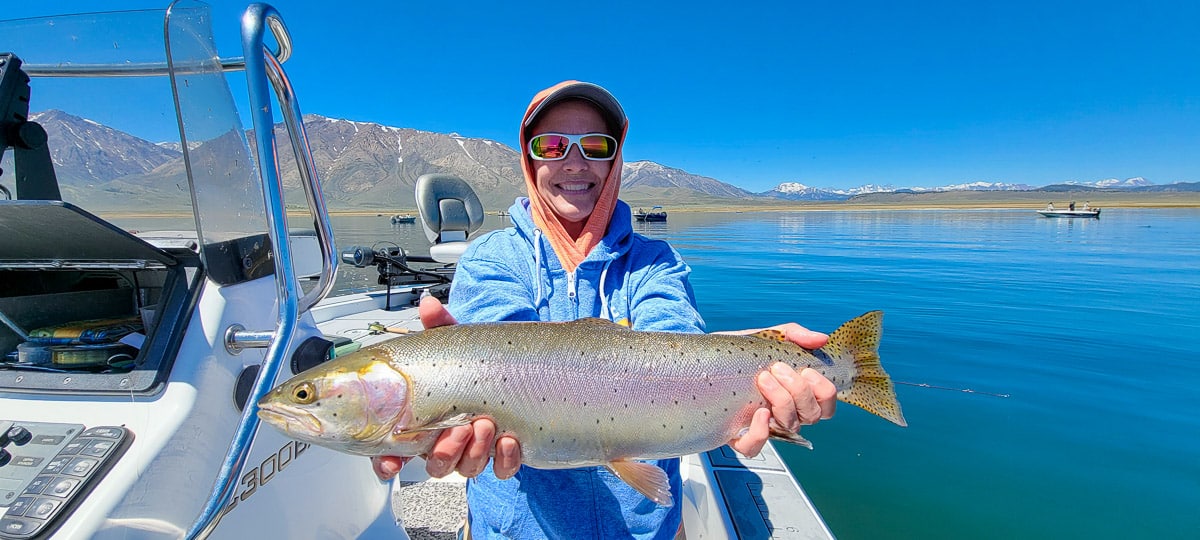 A smiling fly fisherman on a lake in a boat holding a large cutthroat trout.