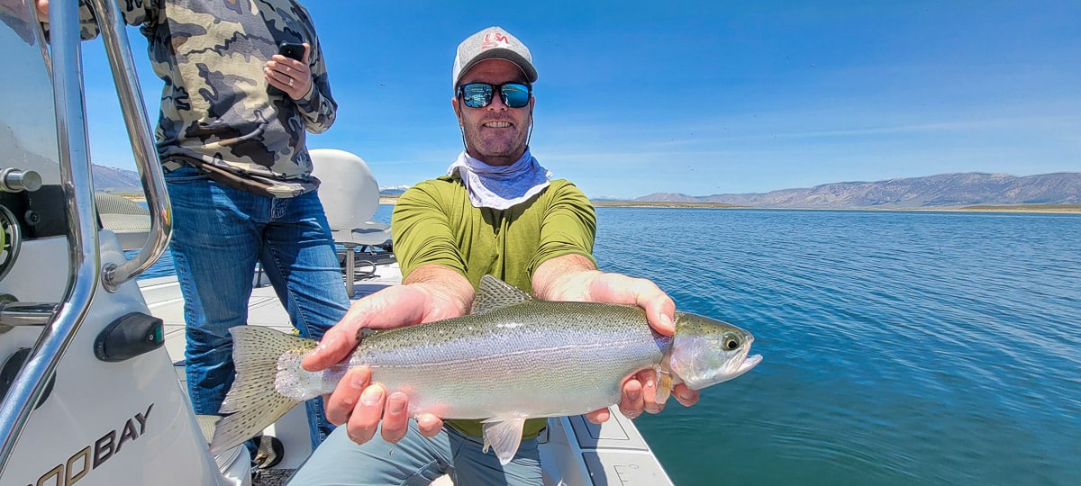 A smiling fly fisherman on a lake in a boat holding a large rainbow trout.