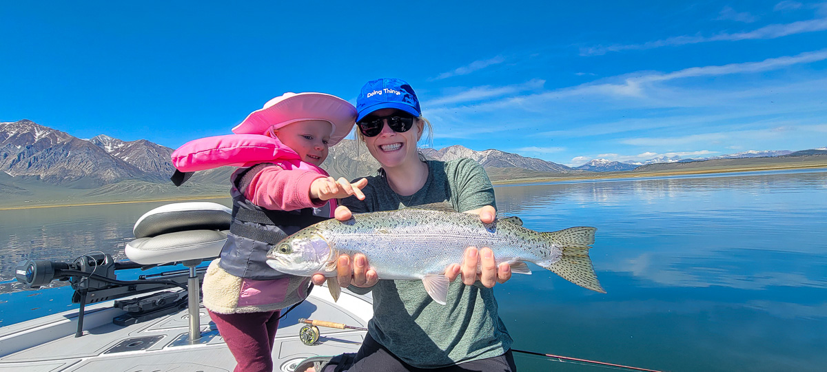 A smiling female fly fisherman and a young girl wearing pink on a lake in a boat holding a large rainbow trout.