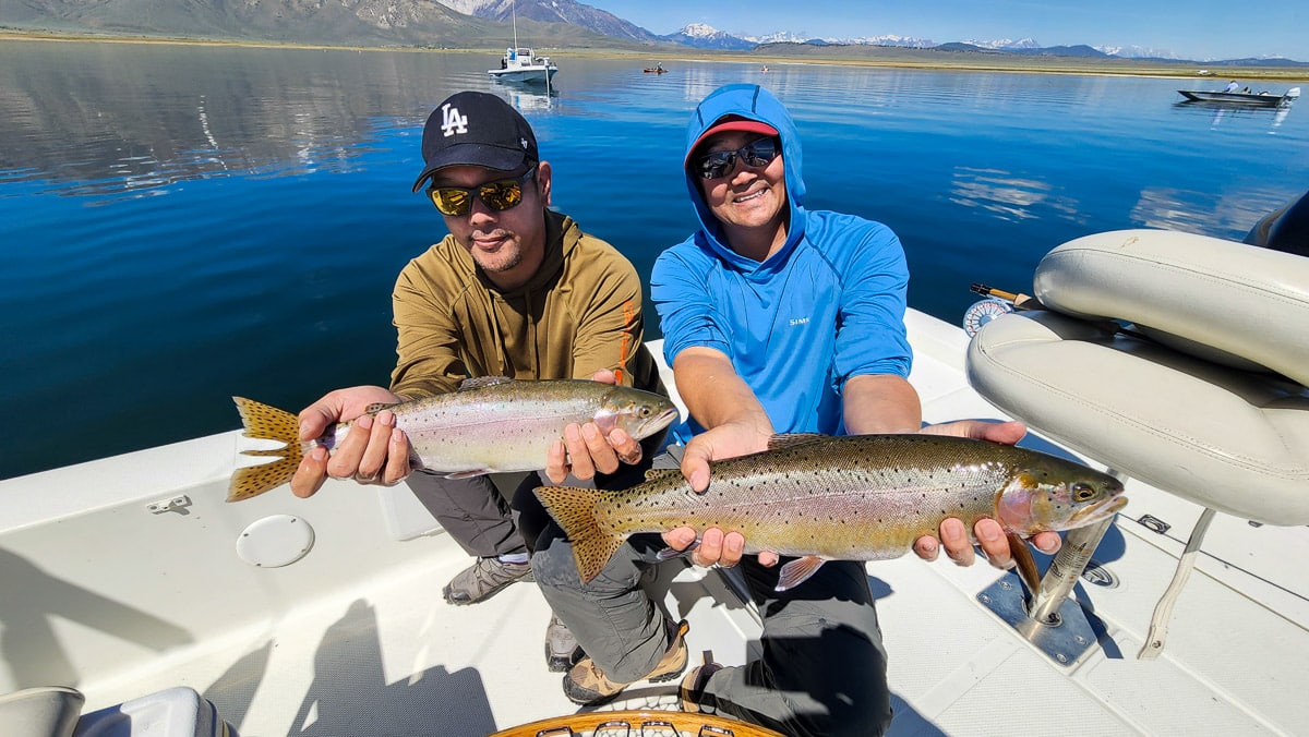 A pair of fly fisherman on a lake in a boat holding 2 behemoth cutthroat trout.