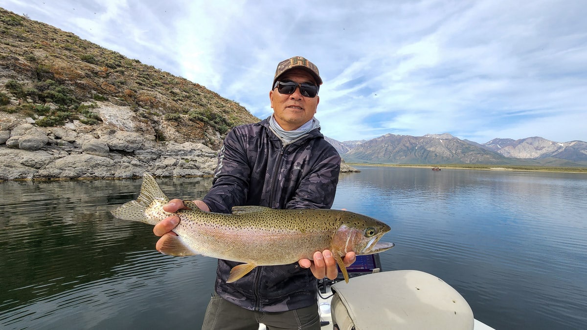 A smiling female fly fisherman and a young girl wearing pink on a lake in a boat holding a large rainbow trout.