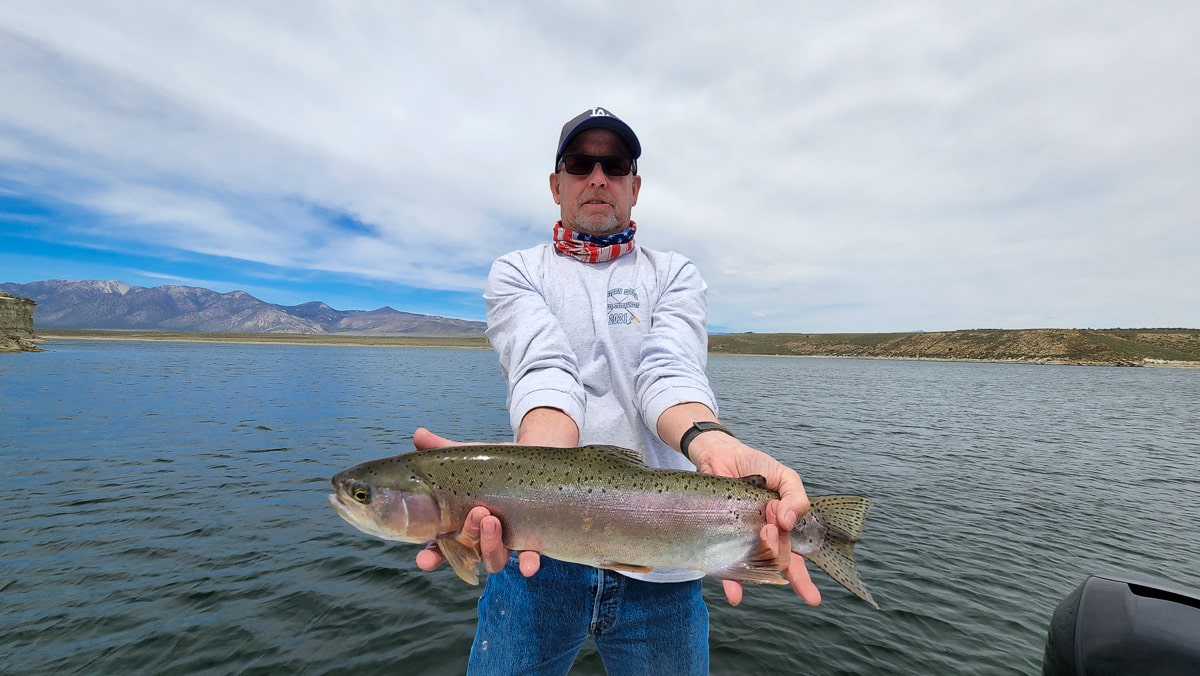 A fly fisherman in a boat holding a large rainbow trout.